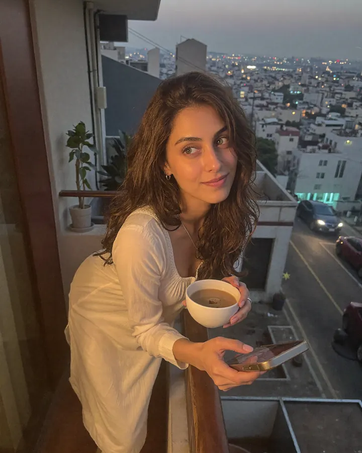 Aisha on a balcony at dusk with a cup of chai, the city lights behind her