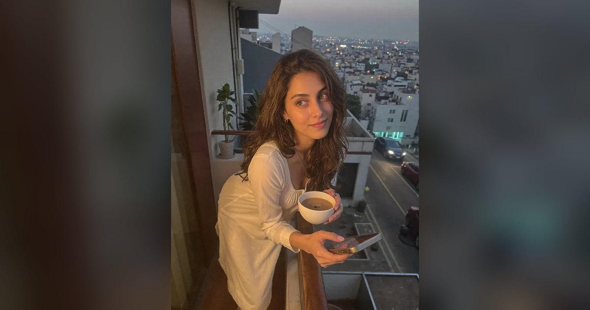 Aisha on a balcony at dusk with a cup of tea and her phone, city lights behind her