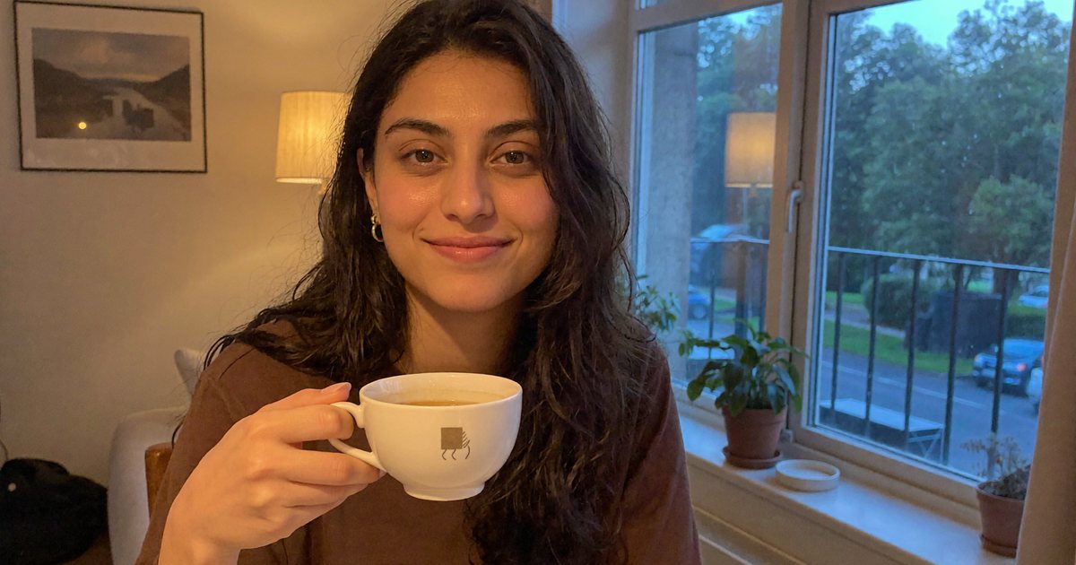 Aisha sitting by a window with a cup of tea in the evening, smiling softly at the camera