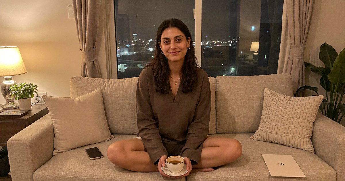 Aisha on a sofa in her living room in the evening, holding a cup of chai
