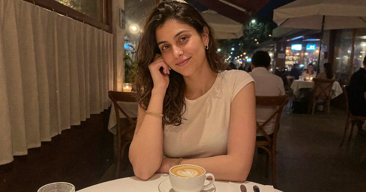 Aisha sitting at a cafe table at night, smiling softly at the camera