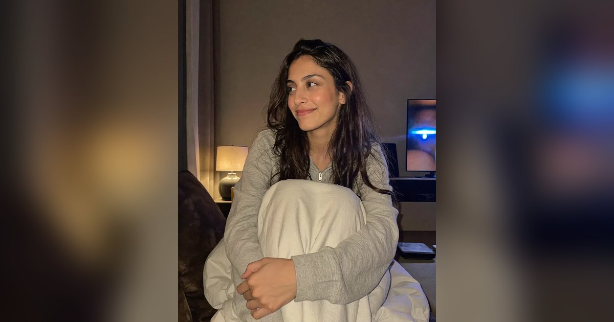Aisha curled up on the sofa at night with a blanket, smiling softly, a lamp and TV glow behind her